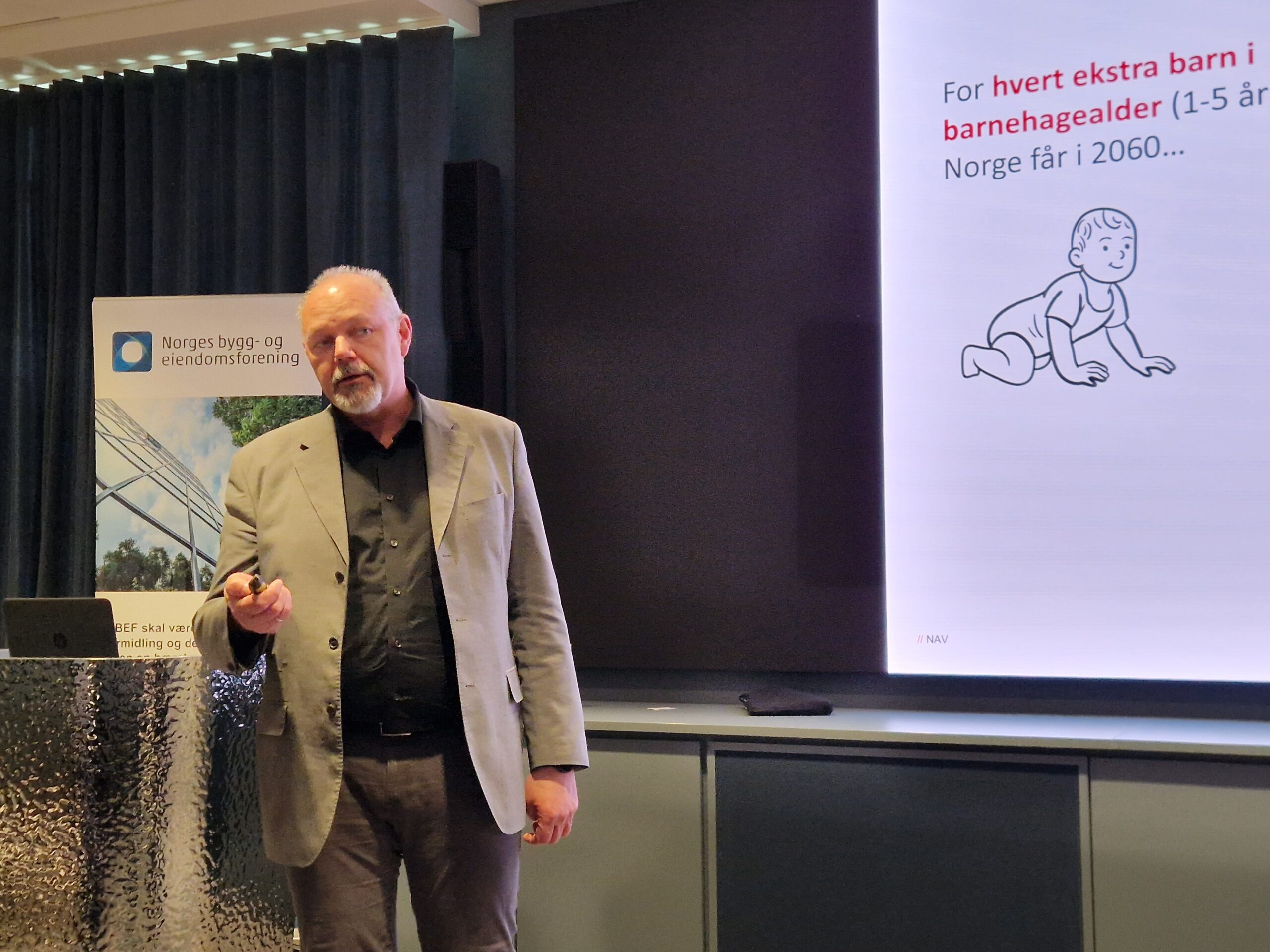 Speaker in a light blazer presenting on a stage beside a banner, with a large slide showing Norwegian text and a baby illustration on the right.