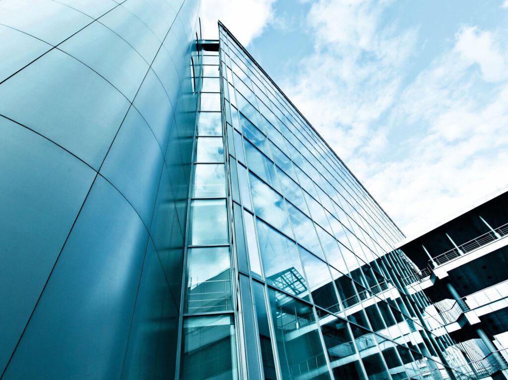 Low-angle view of a blue glass office building with curved panels and metal frames against a bright sky.