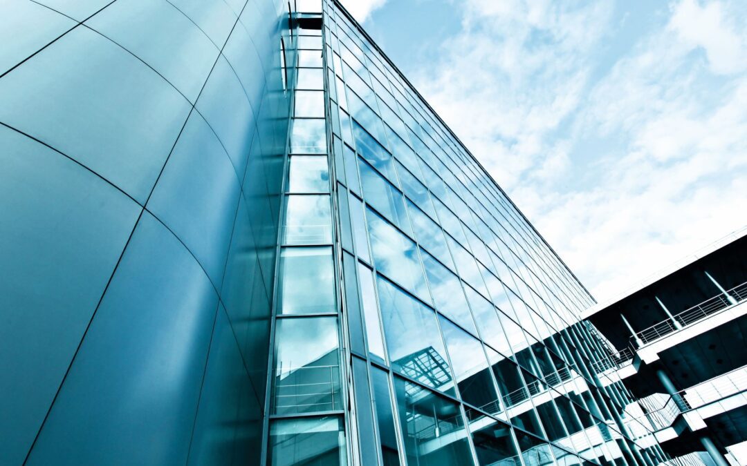 Low-angle view of a blue glass office building with curved panels and metal frames against a bright sky.