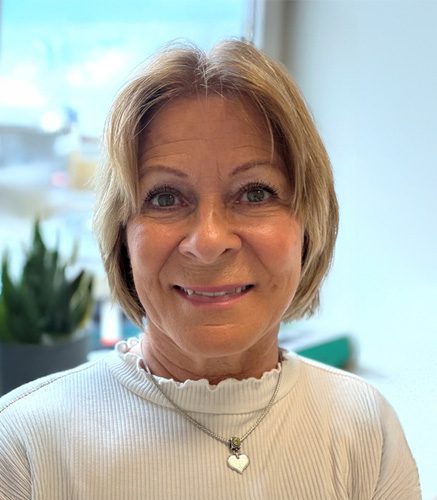 Portrait of a smiling middle-aged woman with short blond hair, wearing a white ribbed top and a heart-shaped pendant necklace.