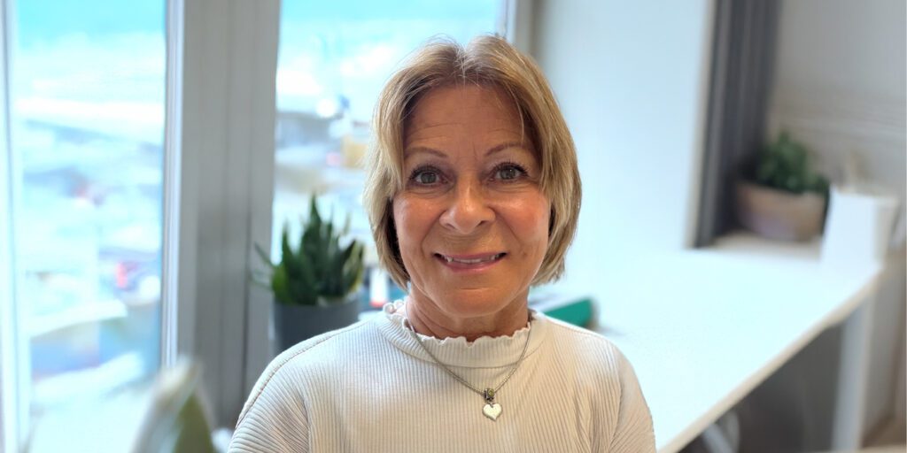 Portrait of a smiling woman with short blond hair, wearing a white ribbed top and heart pendant, in a bright office setting with plants in the background.