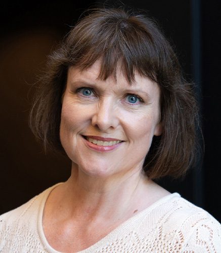 Smiling woman with short brown hair and blue eyes wearing a cream knit sweater, posed against a dark backdrop.