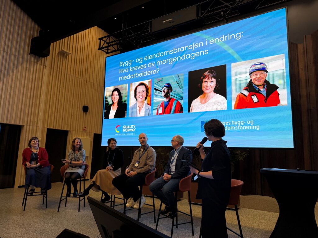 Panel discussion at a conference with seven people seated in a semicircle on stage, microphone in use, large blue screen behind them displaying a presentation slide and slides of portraits at the bottom left.