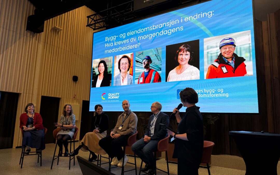 Panel discussion at a conference with seven people seated in a semicircle on stage, microphone in use, large blue screen behind them displaying a presentation slide and slides of portraits at the bottom left.