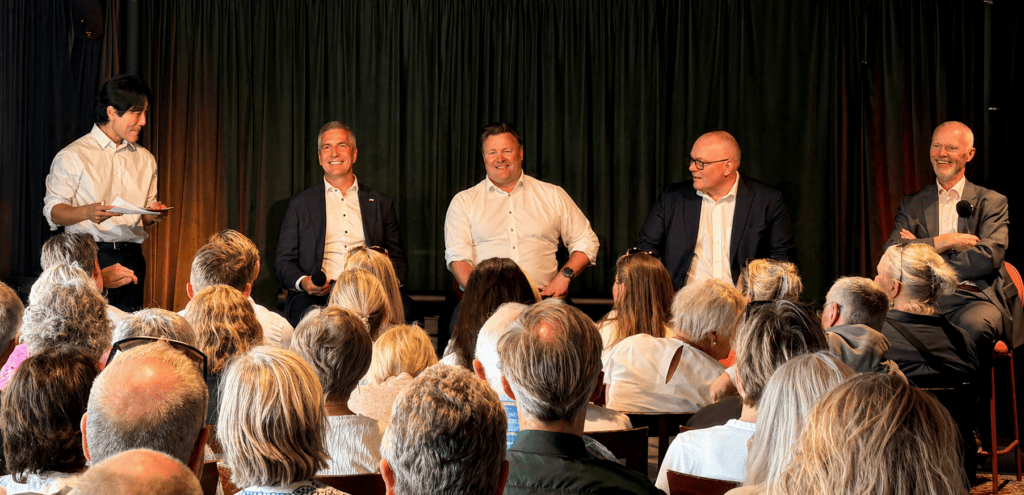 Panel of five men in business attire on a stage, one standing with papers while others hold microphones and listen to the discussion, audience in foreground.
