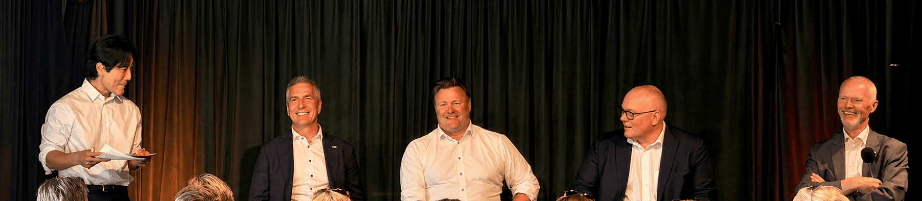 Five men on a stage panel; one standing with papers, others seated and smiling in front of dark curtains with warm stage lighting.