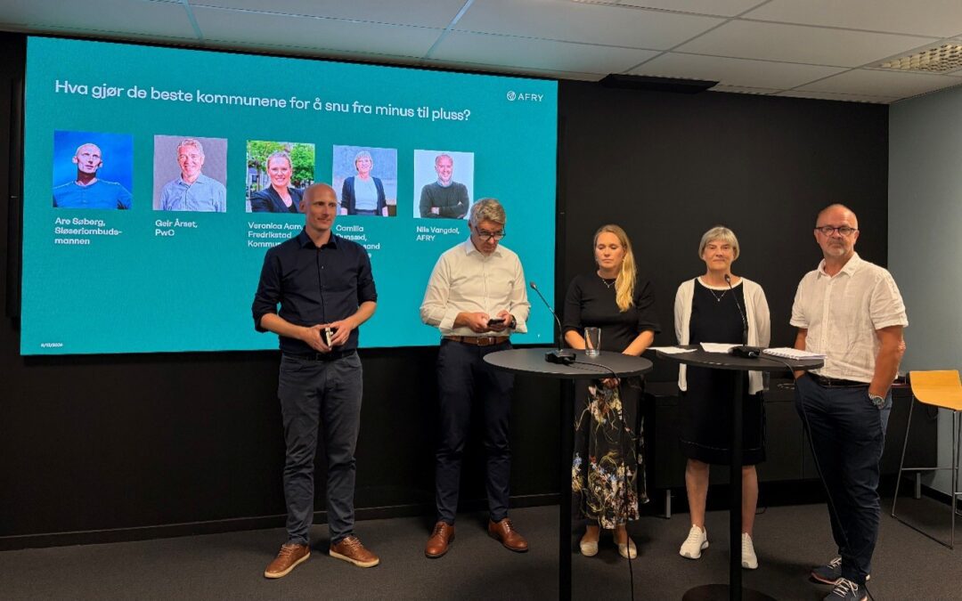 Five presenters stand at a panel in a conference room, a teal slide with portraits and names projected behind them.
